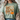 A woman wearing a sage green graphic t-shirt with a fall-themed design that reads 'God is Good ALL THE TIME' alongside a pumpkin and flowers, styled with a beige knit cardigan.