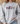 A woman wearing a light gray crewneck sweatshirt with a graphic of four cartoon sports balls (football, basketball, hockey puck, and baseball) walking in a row. The background shows a blurred indoor setting with a woven hat and a sofa.