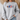 A woman wearing a light gray crewneck sweatshirt with a graphic of four cartoon sports balls (football, basketball, hockey puck, and baseball) walking in a row. The background shows a blurred indoor setting with a woven hat and a sofa.