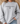 A person wearing a light heather gray crewneck sweatshirt with the black printed text 'the house that built me' and a small house icon on the chest, paired with light blue jeans in a softly lit indoor setting.