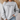 A person wearing a light heather gray crewneck sweatshirt with the black printed text 'the house that built me' and a small house icon on the chest, paired with light blue jeans in a softly lit indoor setting.