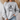 A woman wearing a light gray crewneck sweatshirt with a black graphic of a frog riding a bicycle and the text 'BE HUMBLE BE KIND leave your worries BEHIND'.