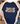 A navy blue graphic t-shirt styled in a flat-lay holiday setting, framed by an open beige knit cardigan. The t-shirt features a distressed graphic reading 'JESUS is the reason' with a patriotic red, white, and blue patterned 'JESUS' and a green patterned 'is the reason'.