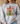 A woman wearing a light gray crewneck sweatshirt with a Christmas graphic on the front. The graphic features a wooden cross with a red and white checkered bow, a green Christmas tree, and the text 'JESUS is the REASON for the SEASON' in red, black, and green lettering.