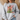 A woman wearing a light gray crewneck sweatshirt with a Christmas graphic on the front. The graphic features a wooden cross with a red and white checkered bow, a green Christmas tree, and the text 'JESUS is the REASON for the SEASON' in red, black, and green lettering.