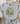 A light gray graphic t-shirt with a tie-front knot, featuring a frog riding a bicycle with flowers and the text 'CHOOSE KINDNESS BE LIKE GOOD Leave you worry BEHIND'. The t-shirt is styled in a flat-lay arrangement with a woven basket, a straw hat, and a beige throw blanket.