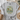 A light gray graphic t-shirt with a tie-front knot, featuring a frog riding a bicycle with flowers and the text 'CHOOSE KINDNESS BE LIKE GOOD Leave you worry BEHIND'. The t-shirt is styled in a flat-lay arrangement with a woven basket, a straw hat, and a beige throw blanket.
