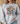 A woman wearing a light gray crewneck sweatshirt with a colorful graphic of a Christmas cross, holiday lights, and the text 'BE THE LIGHT Matthew 5:14'.