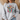 A woman wearing a light gray crewneck sweatshirt with a colorful graphic of a Christmas cross, holiday lights, and the text 'BE THE LIGHT Matthew 5:14'.