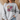 A woman wearing a light heather gray sweatshirt with a colorful graphic of pink and yellow flowers, green leaves, and the text 'CONSIDER the lilies MATTHEW 6:28'.