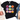 A black short-sleeve graphic T-shirt laid flat on a white background, styled with a cream knit sweater, denim shorts, and beige shoes. The shirt features a grid of nine colorful patterned pumpkins on the front.