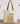 A cream canvas tote bag with brown shoulder straps stands on a wooden surface against a white wall. The front of the bag has a colorful autumn graphic with the text 'HEY THERE PUMPKIN' and an illustration of pumpkins, flowers, and leaves on a light blue background.