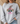 A woman wearing a light gray crewneck sweatshirt with a graphic of a smiling scarecrow face wearing a leopard-print witch hat. The background shows a cozy indoor setting with a straw hat and a couch.
