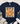 A navy blue short-sleeve t-shirt laid flat on a white wood background, featuring a large autumn turkey graphic with patterned feathers in orange, beige, and brown. The shirt is styled with a fringed cream throw, blue jeans, and white sneakers.