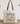 A cream canvas tote bag with brown leather handles, featuring a vintage-style graphic print that reads 'You had me at VINTAGE' above illustrated teacups, flowers, and retro objects. The bag is displayed on a wooden surface against a white wall with eucalyptus branches in the background.